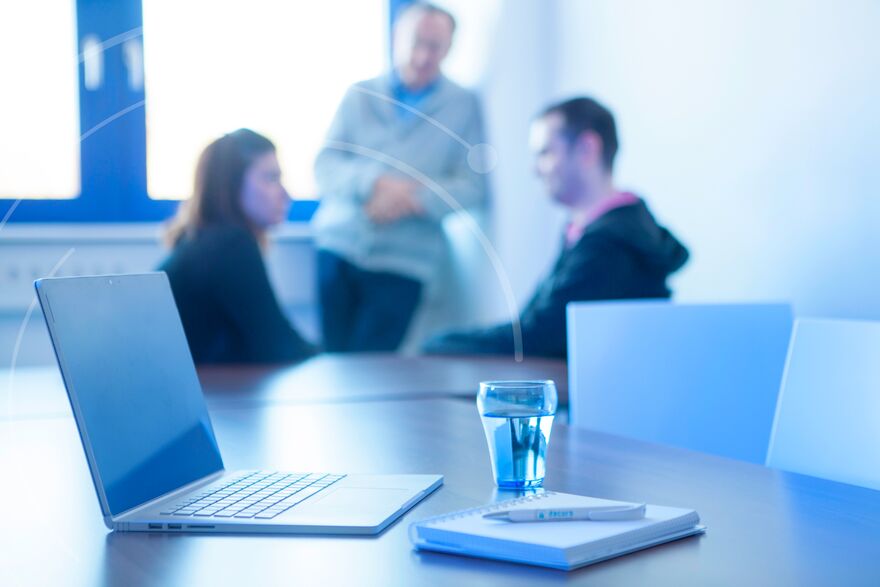 ein aufgeklappter Laptop und im Hintergrund drei Menschen die miteinander reden